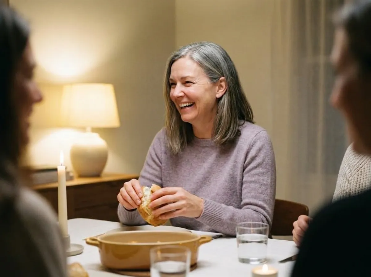 A woman laughing at a dinner table with friends, tearing a crusty bread roll.